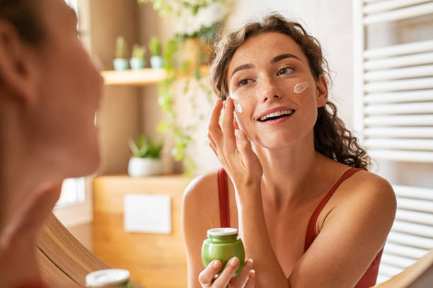 A woman smiling while applying moisturizer in front of a mirror, preparing for her makeup routine.